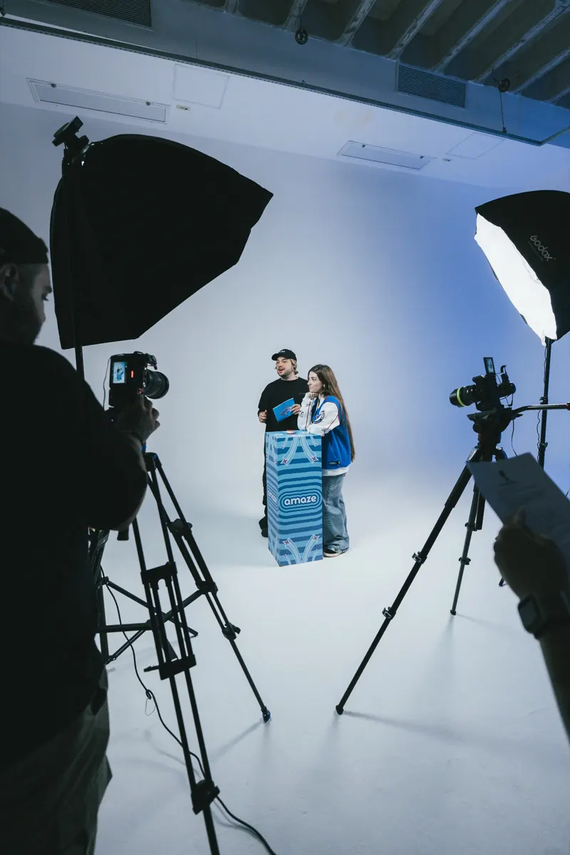 Bastidor do estúdio: câmeras em tripés e softboxes em primeiro plano, e duas pessoas ao fundo, junto a um display azul, sobre fundo branco.