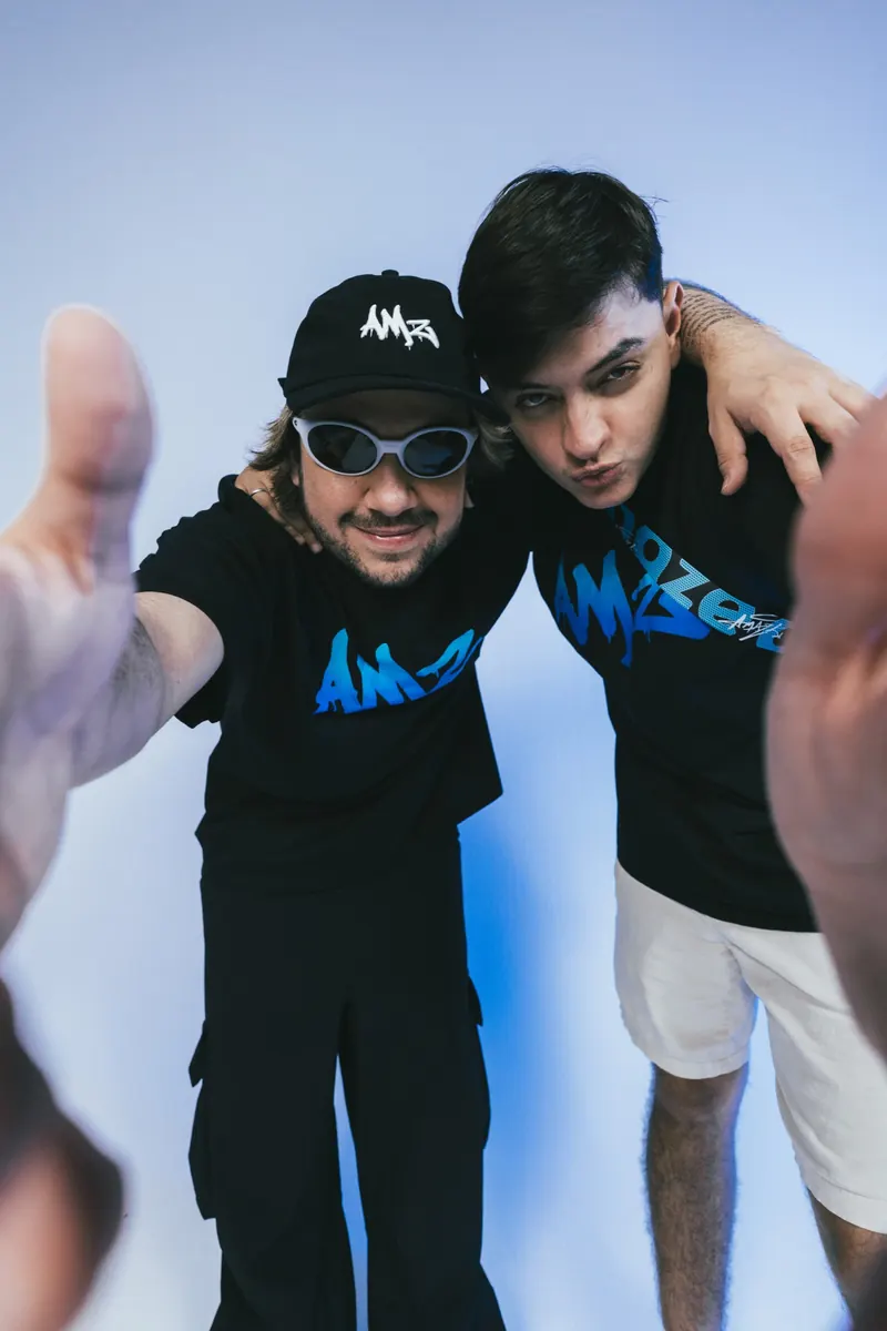 Dois homens de boné e camiseta preta com a marca AMZ, abraçados e inclinados para a câmera, com mãos estendidas em primeiro plano.