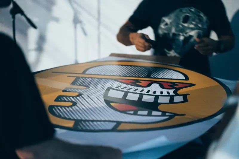 Em primeiro plano desfocado, um adesivo redondo gigante com um rosto estilizado em amarelo e halftone, sobre uma mesa, enquanto uma pessoa de camiseta preta trabalha nele ao fundo.