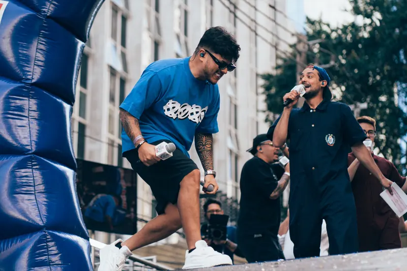 Apresentador de camiseta azul, óculos escuros e microfone na mão se inclina sobre o palco ao lado de outro apresentador, com um arco inflável azul à esquerda.